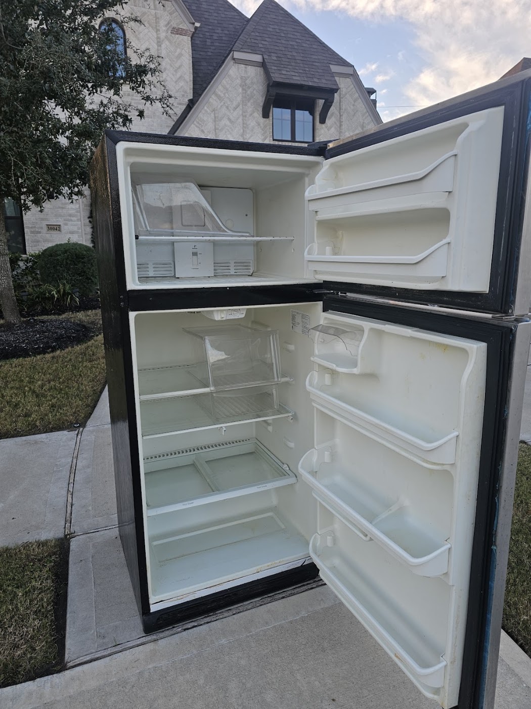 Emptied black top-freezer refrigerator with the doors open outside a home